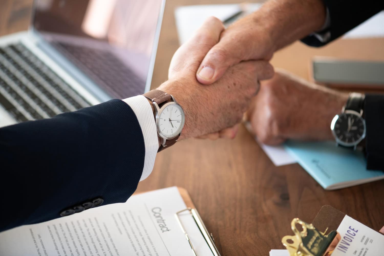 Two professionals shake hands over a wooden desk, with a laptop, papers, and watches visible on the table in a business setting.